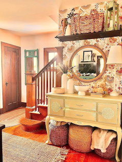Charming cottage room with floral walls, wooden stairs, round mirror, sideboard, and red sofa with patterned pillows