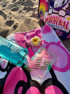 Hello Kity Hawaii towel on sand with turquoise bottle, pink hair clips, and plumeria flower
