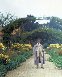 Elderly man in long coat and hat walking on a garden path lined with colorful flowers and green trees