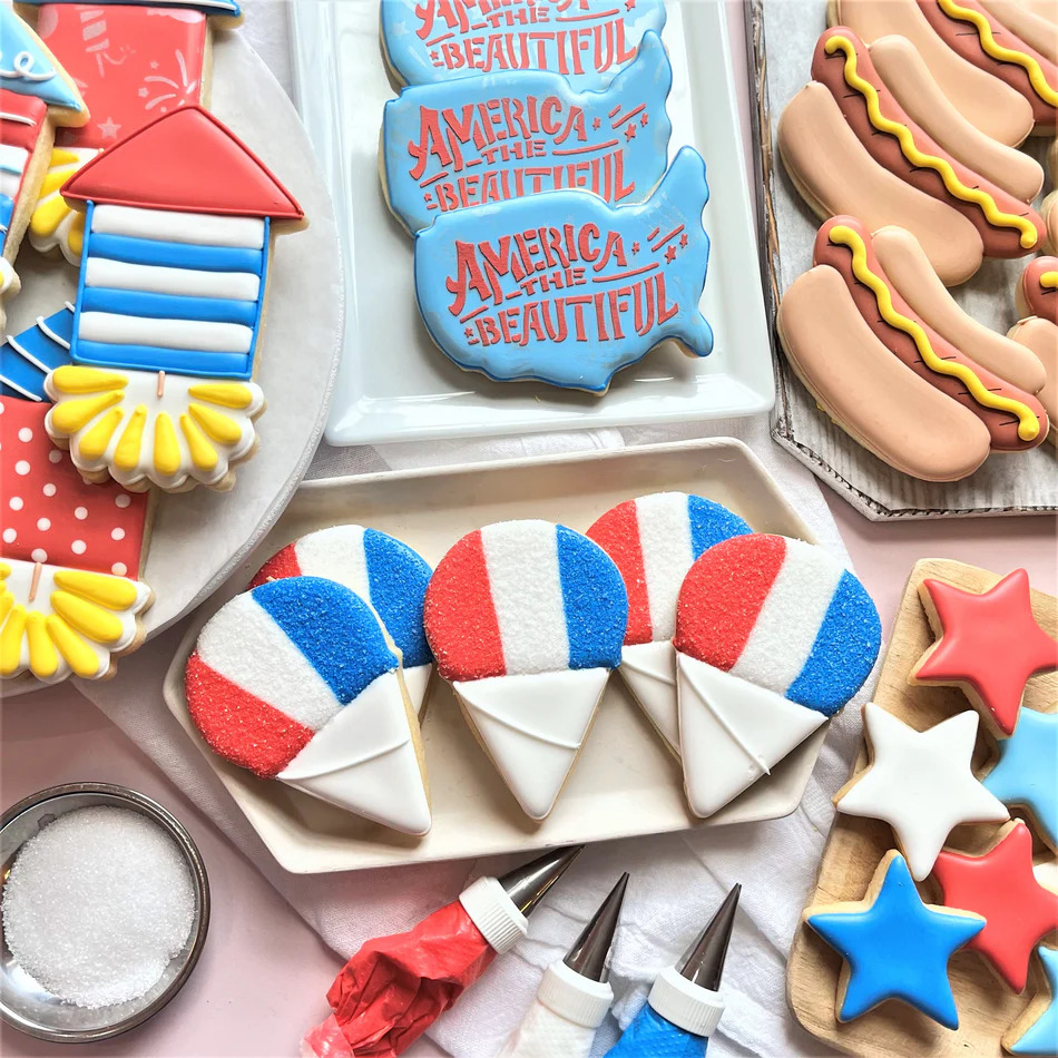 Decorated patriotic sugar cookies in red white and blue with stars, hot dogs, rockets, and America the Beautiful text