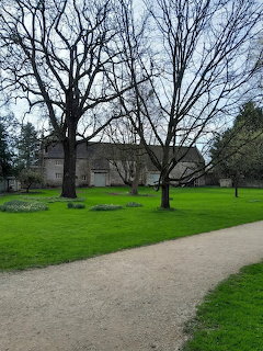 Leafless trees on a green lawn beside a long stone outbuilding and gravel path