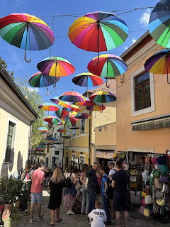Rainbow umbrellas hanging over a busy pedestrian street in Szentendre with crowds of tourists walking below