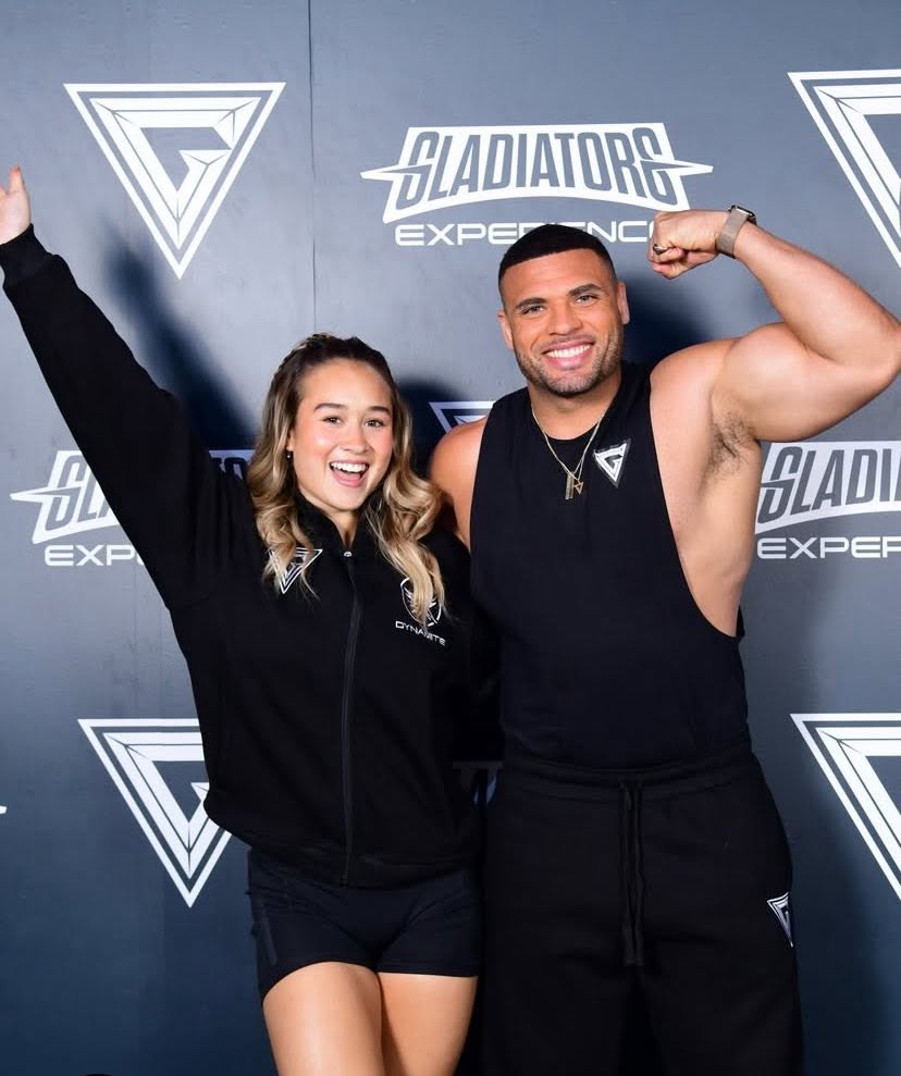 A woman and a muscular man pose together in front of a dark event backdrop