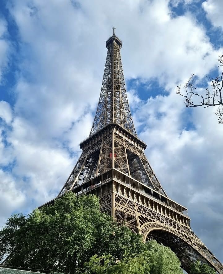 Eiffel Tower viewed from below with trees in front and a cloudy blue sky