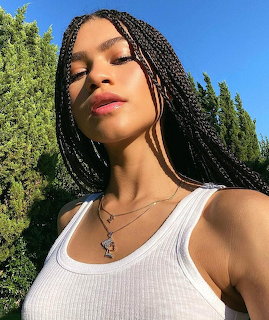 Close-up of a woman with long braids in a white tank top outdoors with trees and blue sky