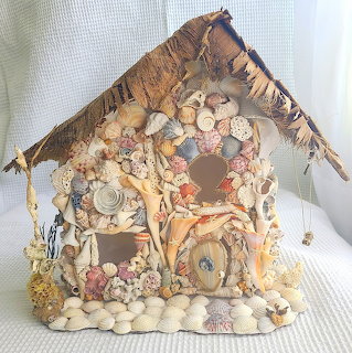 Seashell fairy house craft with shell windows, a shell door, and driftwood roof on white fabric