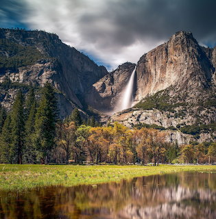 Tall waterfall flowing down steep granite cliffs above a green meadow with pond reflection and autumn trees