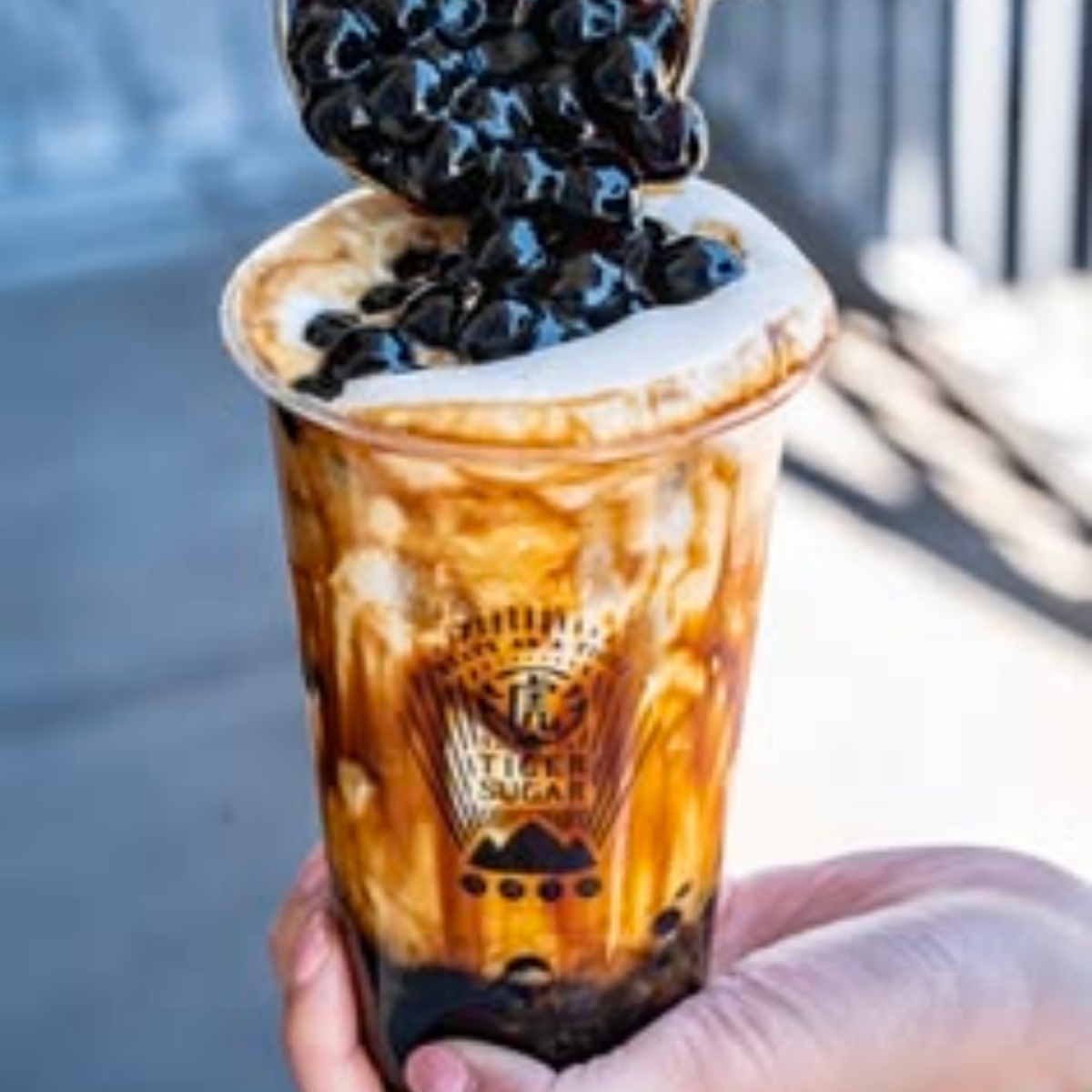 Hand holding iced boba milk tea with tapioca pearls pouring from spoon into cup with brown sugar swirls