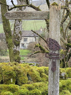Weathered wooden signpost in a mossy garden with a small house behind bare trees