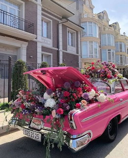 Pink vintage car covered with colorful flower arrangements parked on street with buildings behind