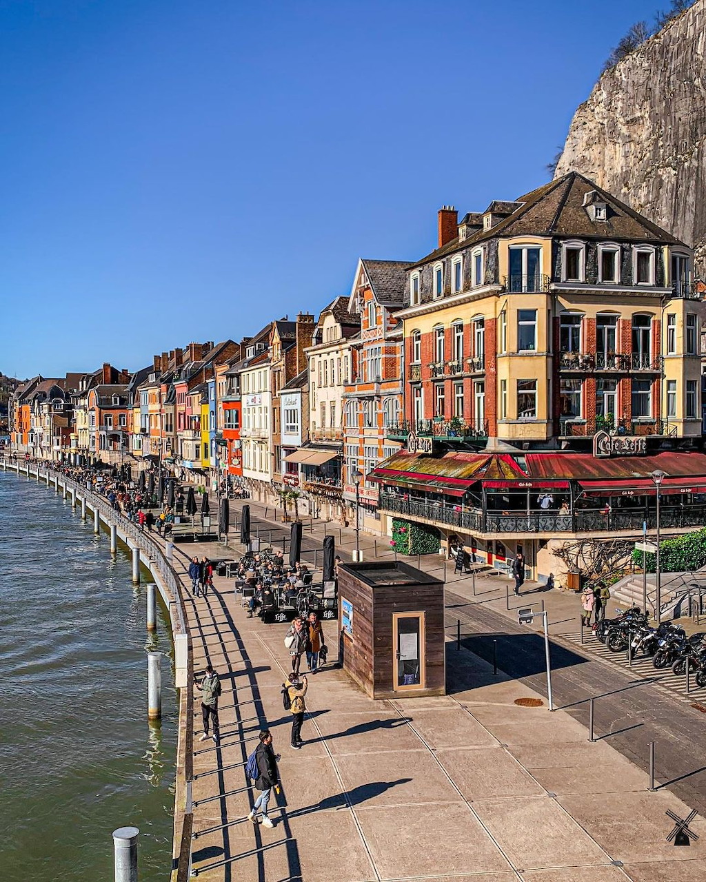 Row of colorful historic buildings along river promenade in Dinant with cliff behind