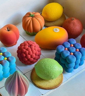 Colorful array of decorative pastries including fruit-shaped cakes, berry tarts, and mousse desserts on white surface