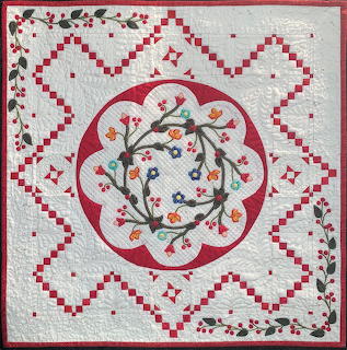 Red and white quilt with geometric borders and central floral appliqué with birds and colorful flowers