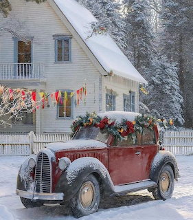 Snow-covered vintage red car with Christmas decorations parked in front of a white house in winter
