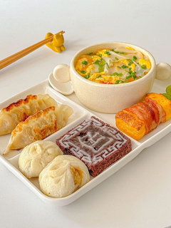 White compartmented tray with Korean dumplings, steamed buns, rice cake, egg roll, and egg drop soup