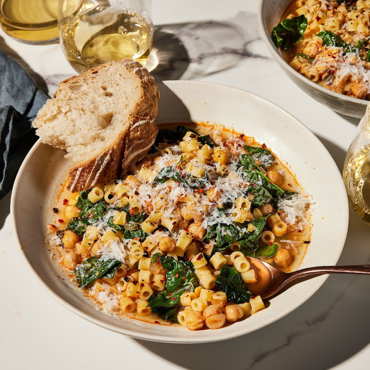 Bowl of pasta with leafy greens, grated cheese, bread, and glasses of white wine