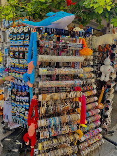 Market stall with jewelry and accessories on rotating display racks under blue canopy with greenery