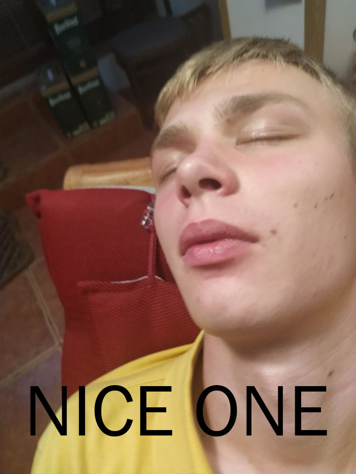 Close-up of a young blonde man with eyes closed, resting on a red cushion, with text 'NICE ONE' overlaid