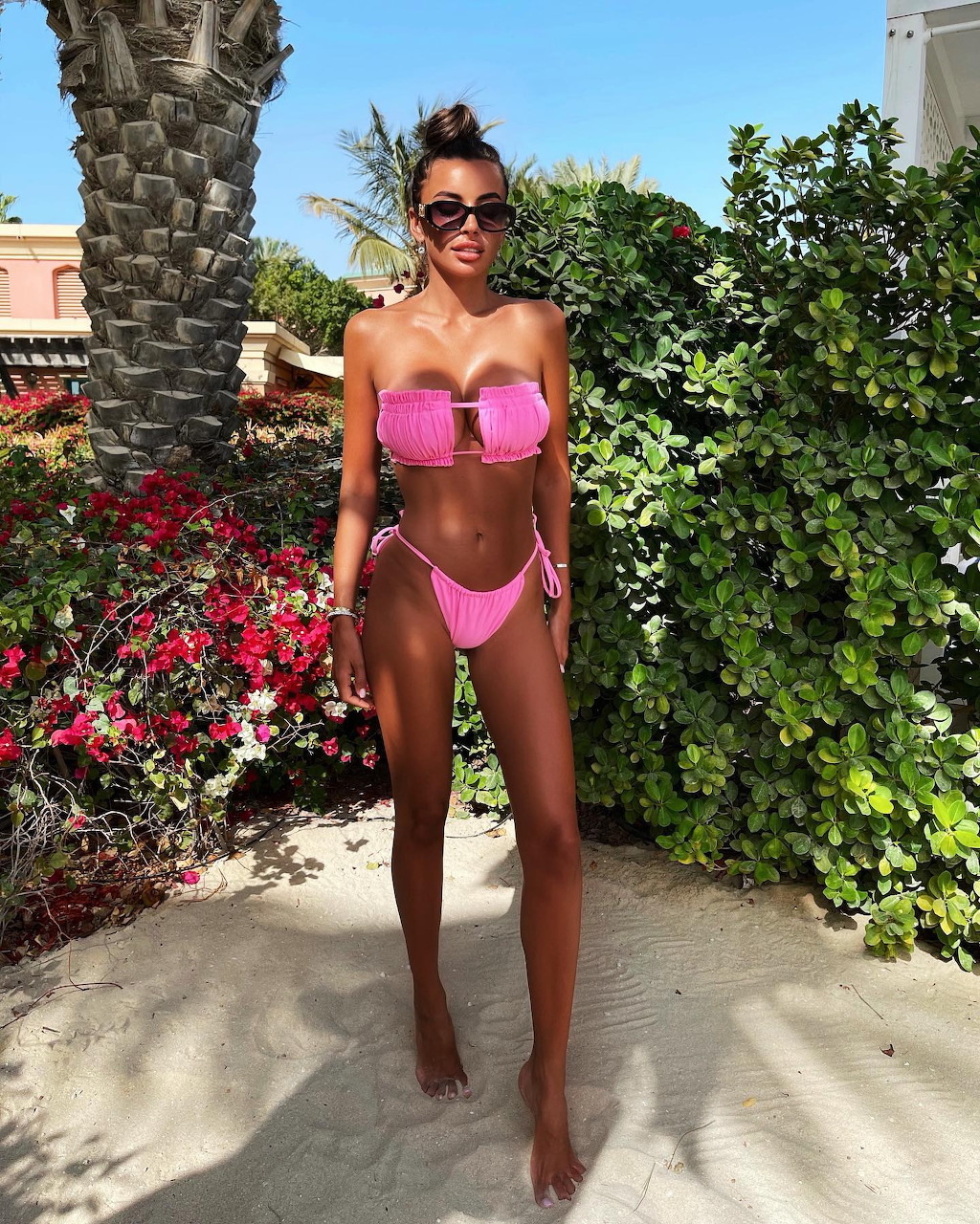 Tanned woman in pink bikini standing on sand with tropical plants and palm tree behind her