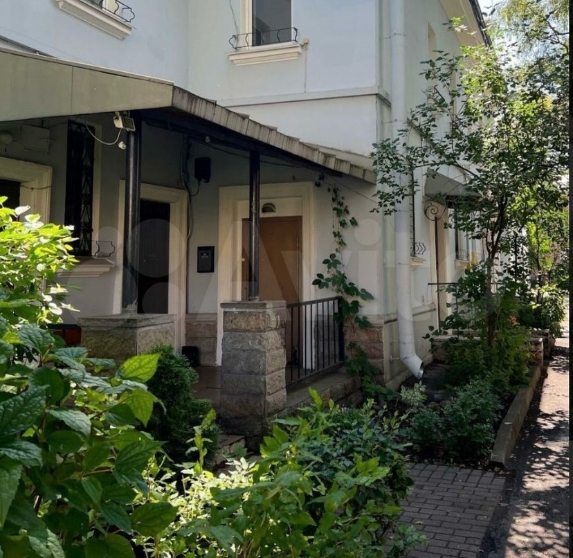 White cottage entrance with dark awning, brick path, and surrounding garden plants in sunlight