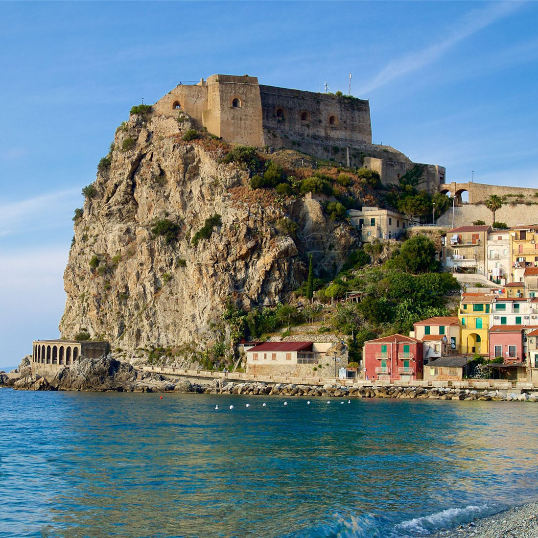 Rocky cliff with ancient fortress above colorful coastal town buildings along calm blue water