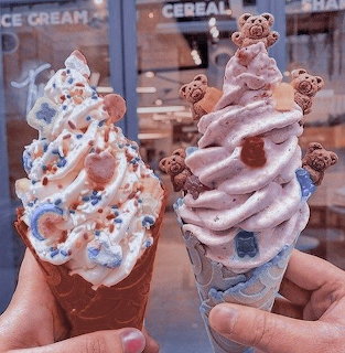 Two hands holding decorated soft-serve ice cream cones with bear toppers in front of a shop window