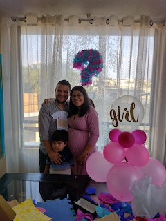 Family of three smiling at a gender reveal party with pink balloons and a girl sign
