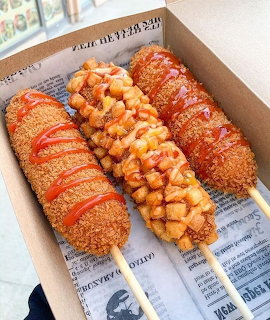 Four Korean corn dogs on sticks in a box with various toppings and sauces
