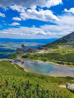 Alpine lake in High Tatras mountains with stone shelter, green meadows and expansive valley below blue sky