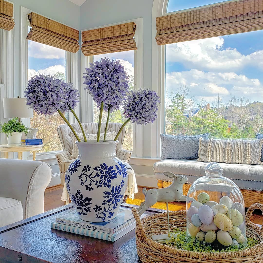 Sunroom with purple allium flowers in blue and white vase, Easter eggs basket, white furniture, bamboo shades, garden view