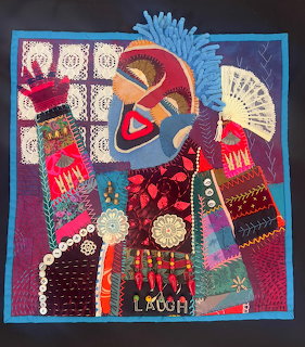 Colorful textile collage art with African mask, fabric patches, beads, lace, and embroidery on dark background