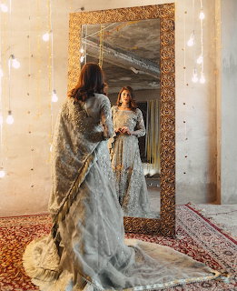 Woman in embellished gray gown viewing her reflection in an ornate framed mirror with string lights