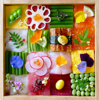 Colorful mosaic grid of sushi pieces, vegetables, and decorative food arranged in a square wooden frame