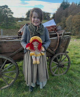 Girl in historical dress holding doll beside old wooden wagon outdoors