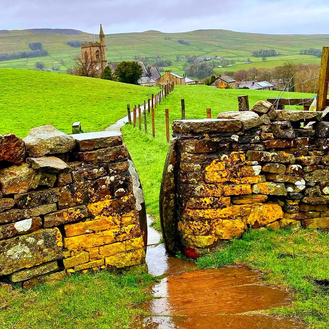 Stone walls with orange lichen lead to church spire and green hills in Hawes, North Yorkshire countryside