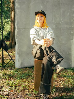 Woman with orange hair in black cap and grey hoodie sitting outdoors against concrete wall