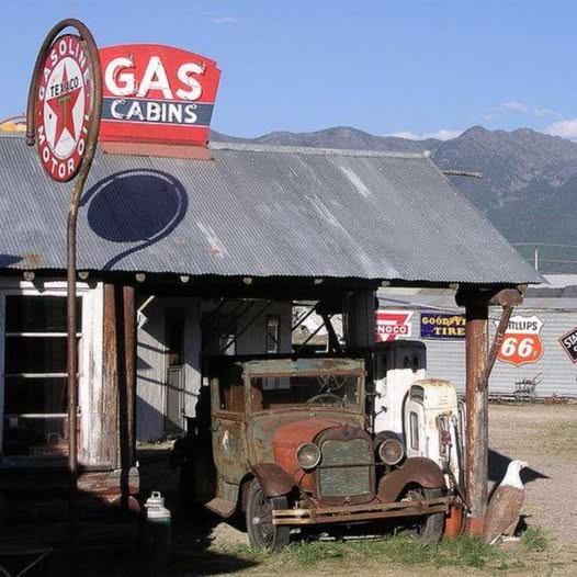 Rusty vintage car parked under an old gas station canopy with retro signs and mountains behind
