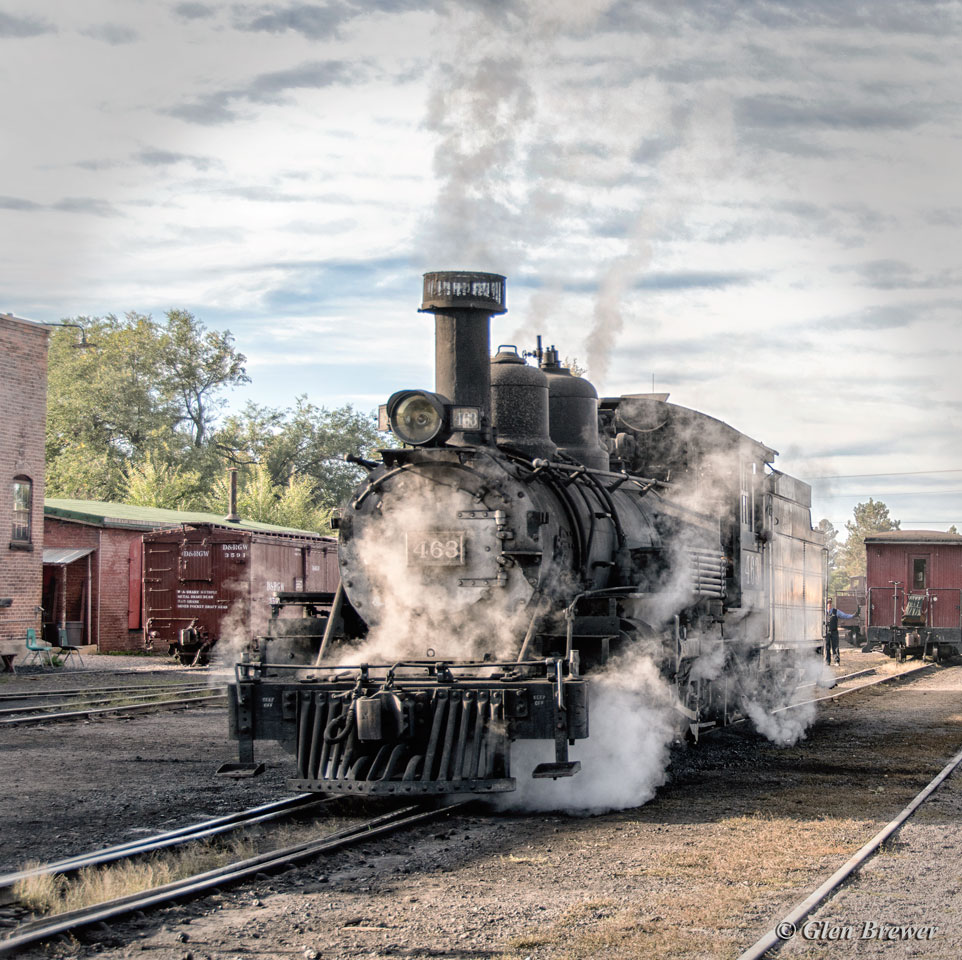 Black steam locomotive with white steam clouds at railroad yard with brick buildings