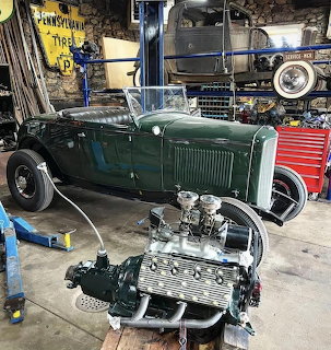 Green classic hot rod car being restored in cluttered garage workshop with engine in foreground