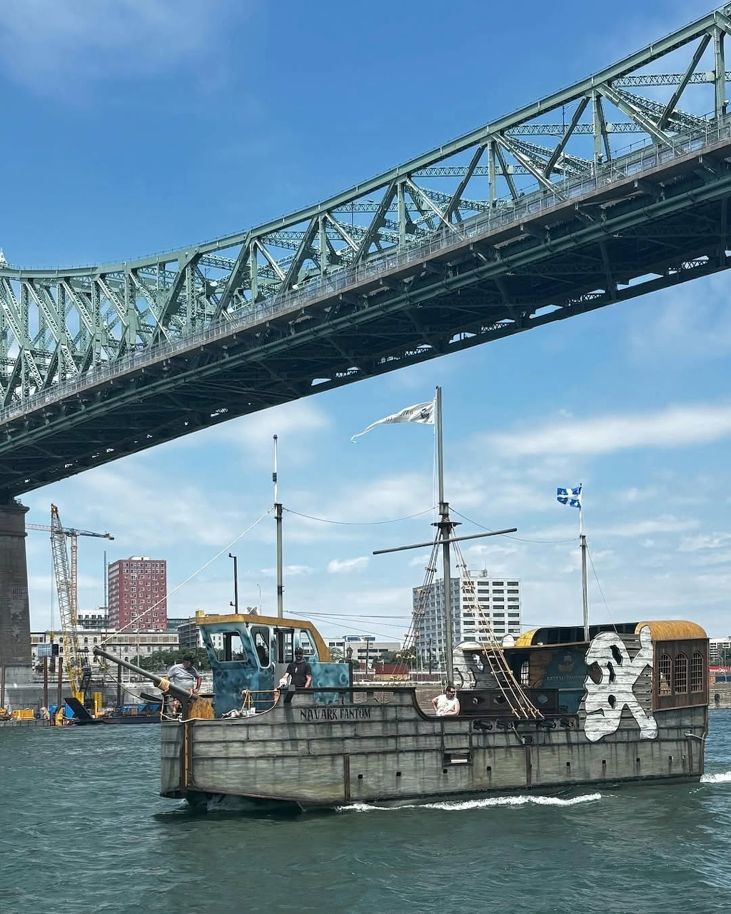 Small weathered boat on the water beneath a large steel bridge with city buildings behind it.