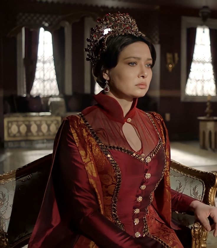 Woman in a red historical gown and jeweled crown seated in an ornate interior