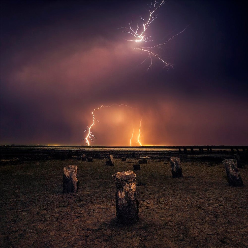 Multiple lightning bolts striking over a desert landscape with dead tree stumps under stormy clouds