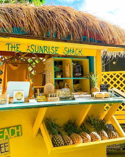Bright yellow Sunrise Shack tropical stand with thatched roof, pineapples, and beverages on display