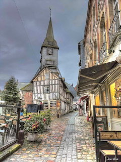 Coblestone street in Honfleur with half-timbered buildings and a church steeple under grey skies