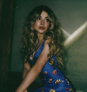 Young woman with long curly blonde hair in a blue floral dress posing in dim lighting