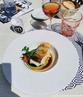 Seared cod fillet on a white plate with vegetables and sauce, rosé wine glasses in background