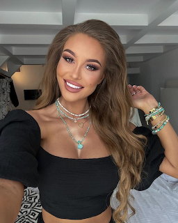 Smiling young woman with long brown hair, black crop top, layered necklaces and bracelets taking a selfie