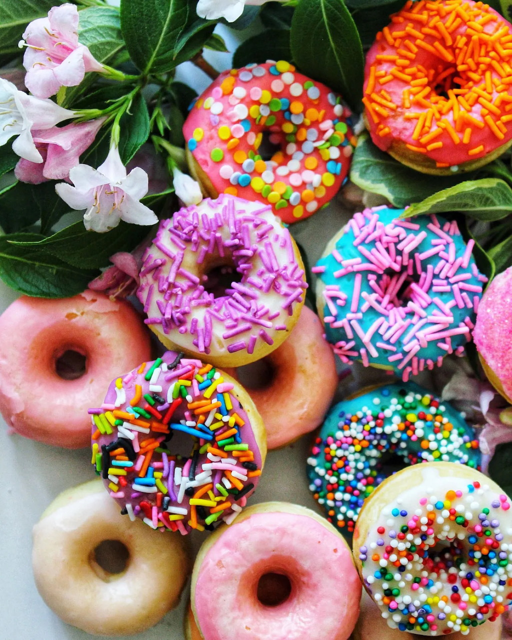 Assorted colorful donuts with frosting and sprinkles beside pink flowers and green leaves