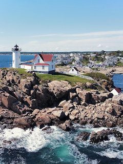 White lighthouse with red roof on rocky cliff surrounded by ocean waves and coastal town in background