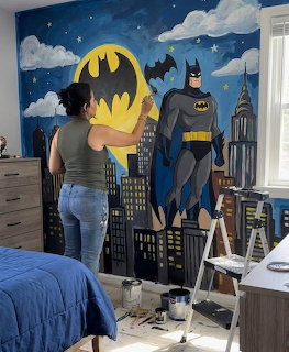Woman painting a Batman mural with cityscape and bat signal on a bedroom wall